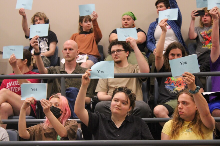 A group of people seated in an auditorium hold up blue cards that read "Yes." Most people appear attentive and engaged, with cards raised above their heads, participating in a vote or decision-making process.