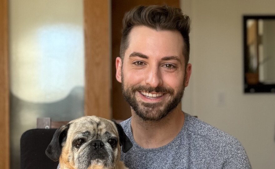 A smiling man with short dark hair and a dark beard is wearing a grey sweater and holding a black and tan pug.
