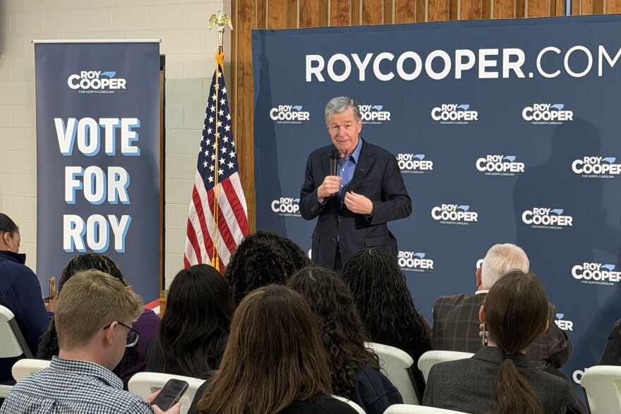 Man in suit with no tie holds microphone in front of campaign banner and before small crowd in room