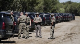 A group of officers stand on a dirt road behind a line of SUVs.