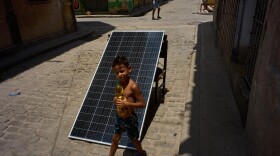 A child walks with a bottle of oil past a solar panel set up on the street to charge batteries during a blackout in Havana, Cuba, Monday, July 6, 2026.
