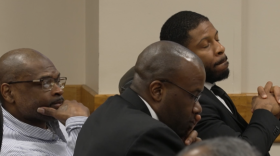 Three men sit in a courtroom, listening during proceedings.