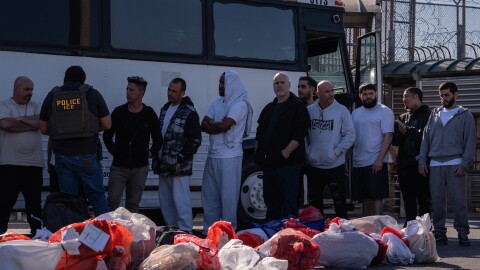 A number or men line up next to a bus while a man with a 'Police ICE' vest speaks to some of them. 
