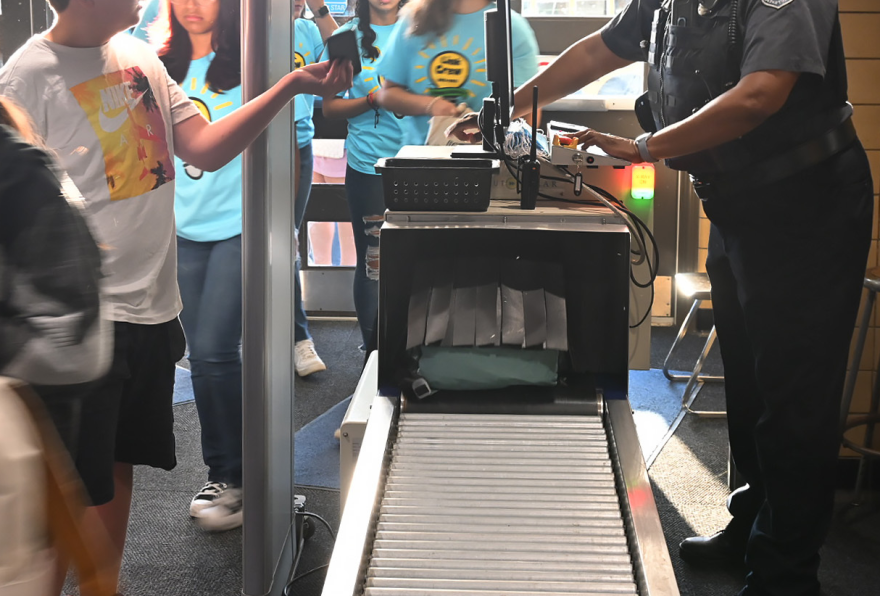 Interior photo shows a group of students lined up entering a building past a security officer and through a metal detector. Also in the picture is a conveyor belt that is carrying backpacks through an X-ray machine.
