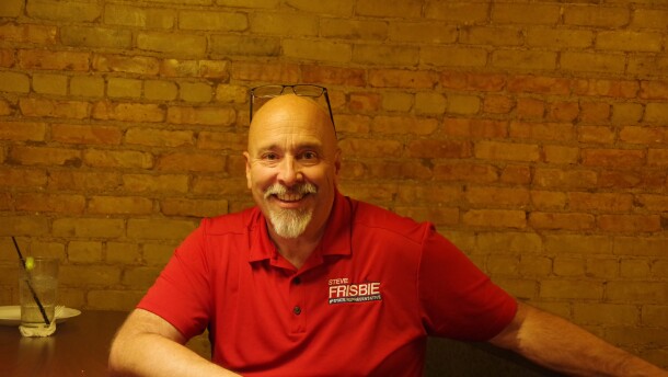 Steve Frisbee in a red campaign logo polo shirt sits at a table with his left arm extended onto a railing.  A campaign sign is on the table along with a cell phone and a clear liquid drink with a black straw.