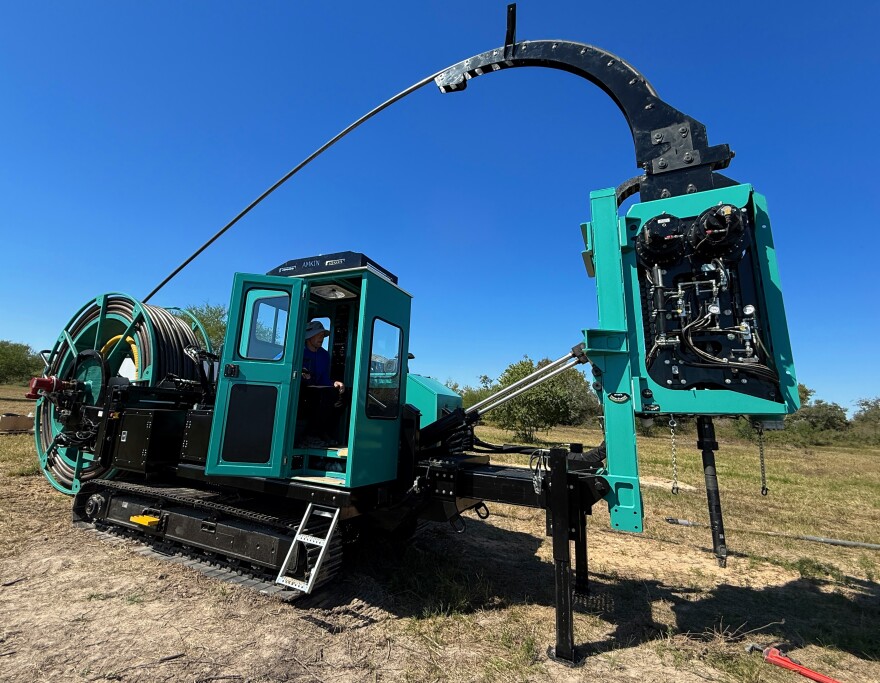 A man sits in the cab of a big, teal piece of heavy machinery on treads, with a giant spool in the back that feeds into a drill bit on the front.