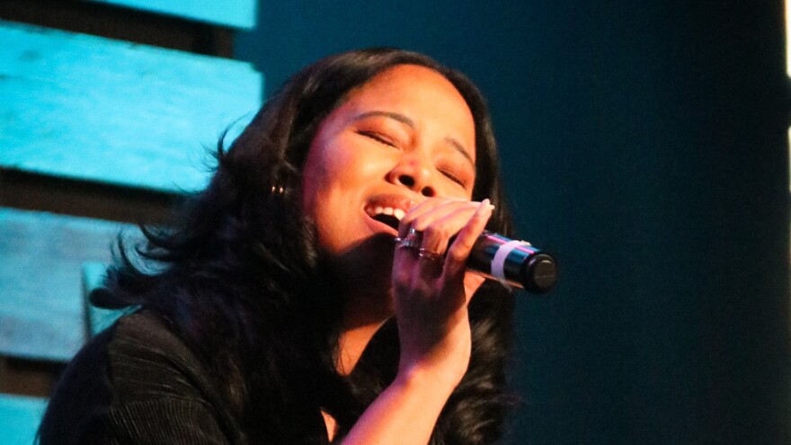  A woman sings into a handheld microphone with her eyes closed in a darkened performance space.