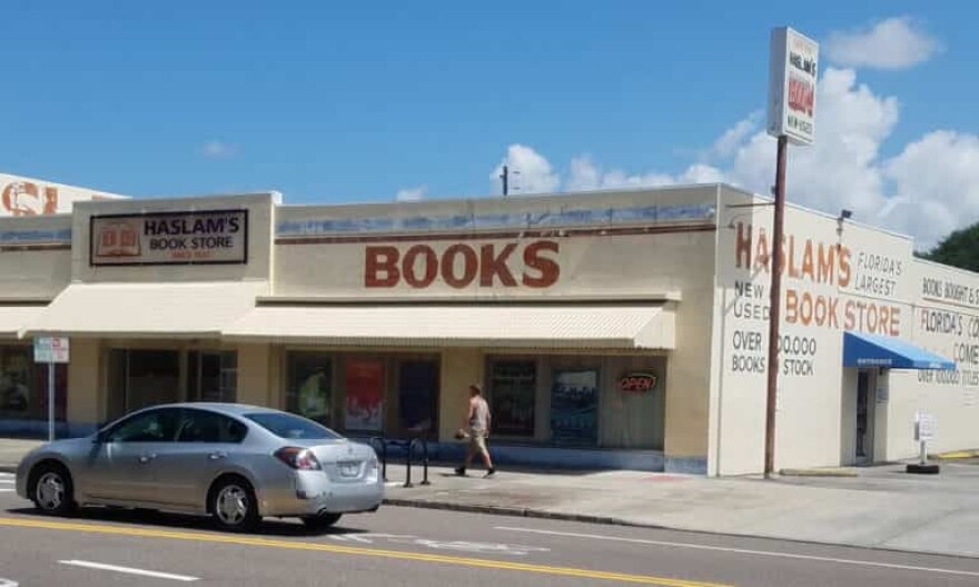 A light brown building that says Haslam's Book Store at the top and Books in large red lettering is seen from across a street with a man walking in front of it and a gray car passing to the left