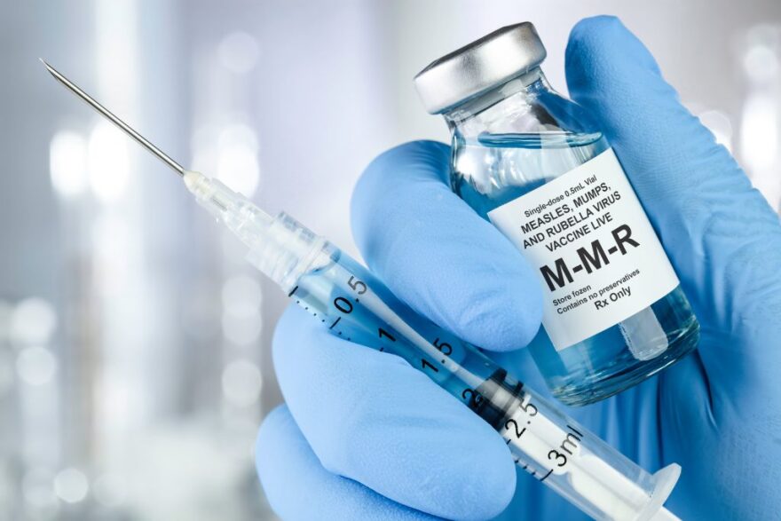 syringe and bottle of vaccine labeled MMR held by a hand wearing a medical blue latex glove 