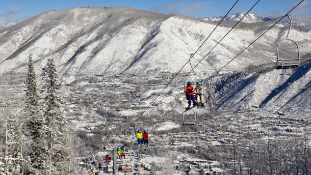 Skiers and snowboarders ride up a ski lift with snowy hills and a town in the background.
