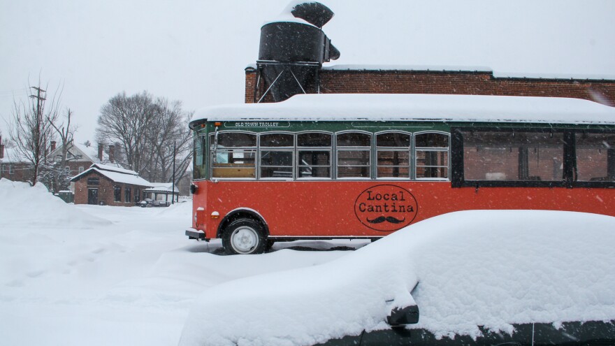 Snow piles up on a trolley parked in front of a brick building. The Local Cantina logo is painted on the side of the trolley. 