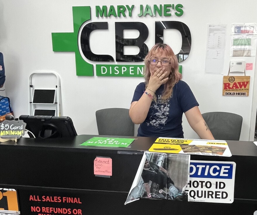 A young woman with pink highlights stands behind a counter that says "Notice: Photo ID Required." A sign behind her reads "Mary Jane CBD Dispensary."