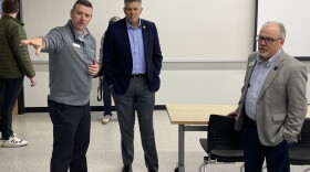 Three men standing in a room with the man on the right pointing with his right arm and the two others looking ahead with their hands in their pockets and a white wall and whiteboard behind them.