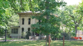 An off-white two-story building is obscured by trees which populate much of the image. Most of the trees stand taller than the image can capture, with branches dangling into the the frame. A road in front of the building is surrounded by metal fences, blocking off access to the property.