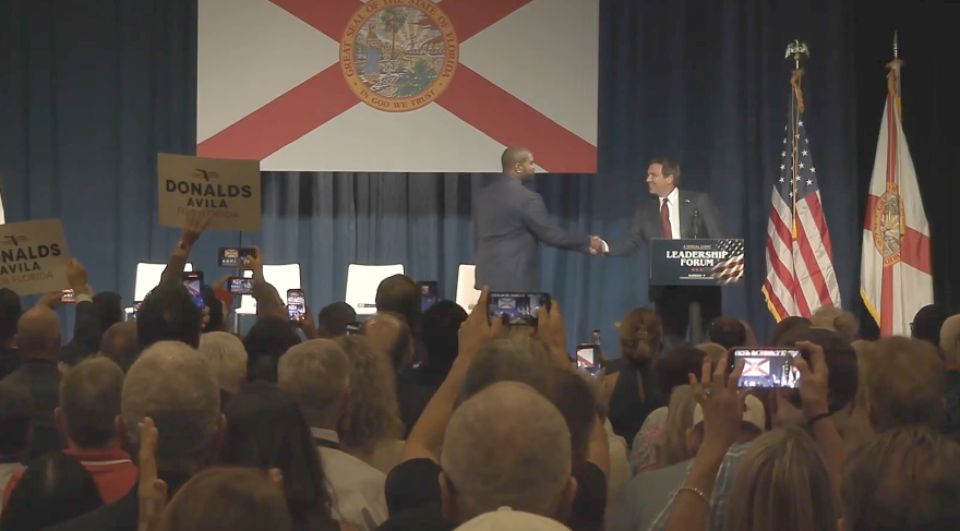 Florida Gov. Ron DeSantis shakes hands with U.S. Rep. Byron Donalds onstage in front of a large Florida flag as audience members watch and take photos.
