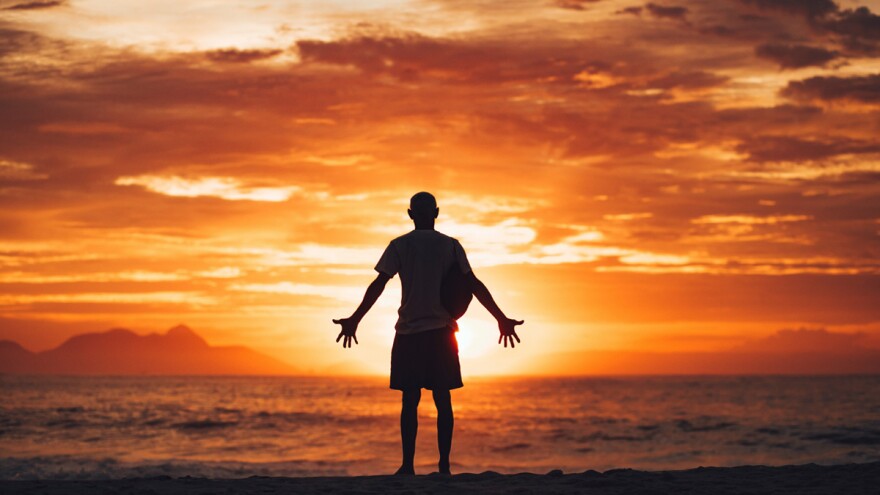 Person standing on a beach at sunset with arms outstretched, facing a vibrant sky over the ocean.
