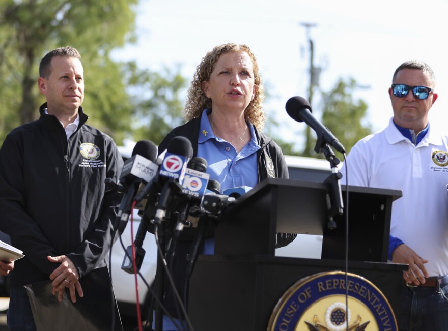 Woman with curly blonde hair speaking at a podium with a man on either side