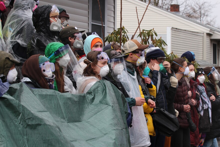 Protesters in masks and goggles stand in a clump, with their arms linked, outside of the entrance to a home.