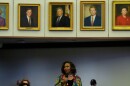 Black woman at the bottom of the photo speaking into a microphone with five portraits in gold frames on a wall above her