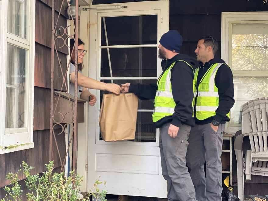 Two village employees wearing reflective vests stand on a porch and hand a brown paper bag to a woman opening a front door.