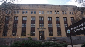 The tan limestone Michigan Avenue Courthouse with Kalamazoo County Building carved at the top of the building. Two trees without leaves to the left and right of the building and a green bush in front of the grey concrete stairs that lead up to the building. A brown sign to the right of the bush reads Michigan Avenue Courthouse.  