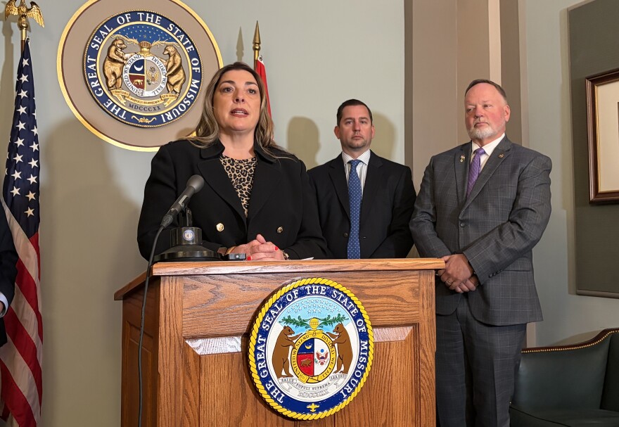 A woman in a black blazer and leopard print shirt speak at a podium with a microphone. The front of the podium has the seal of the state of Missouri on it. Two men in suits stand behind her. 