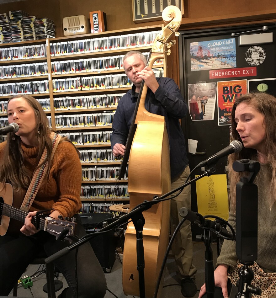 Two seated women play guitar and sing into microphones in a radio studio. Behind them a man plays stand-up bass. Behind all of them is a wall of CDs on shelves.