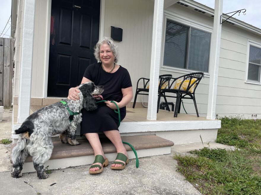 Woman with shoulder-length gray hair wearing a black dress and green sandals sitting on her front porch with her black and white dog.