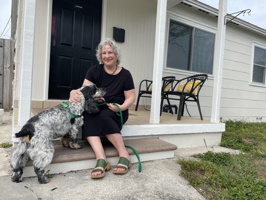 Woman with shoulder-length gray hair wearing a black dress and green sandals sitting on her front porch with her black and white dog. 