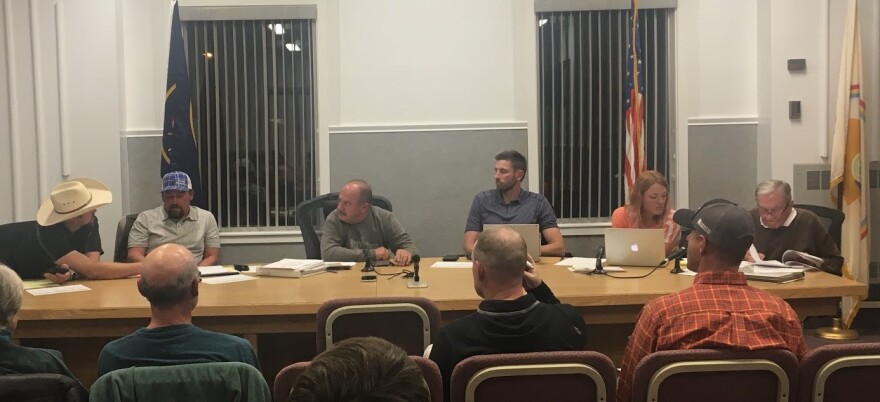 Photo of six people seated at a conference table, as an audience seated in chairs looks on. 