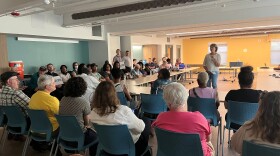 There's a woman addressing a crowd of people seated behind tables in a horseshoe formation. 