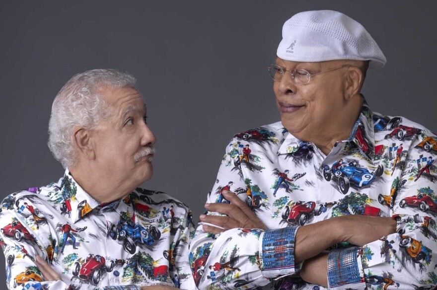 A shorter man with white hair and a mustache looks back at a taller man wearing glasses and a backwards white cap. They are in matching shirts covered with colorful race cars.