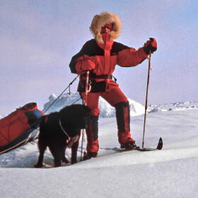 A person standing on skis on a snowy landscape, pulling a red bag behind them. There is a black dog.