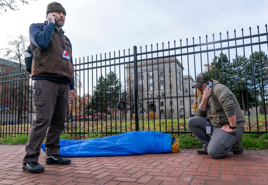 Outreach worker Ryan Hazlett, with Mental Health & Addiction Association of Oregon, and a representative from Recovery Works NW, right, make calls to find treatment options for a man sick from fentanyl, center, in downtown Portland, Ore. on Dec. 13, 2023. The work was part of a pilot program by Portland Police Bureau’s bike squad and addiction recovery providers that launched in December 2023. 