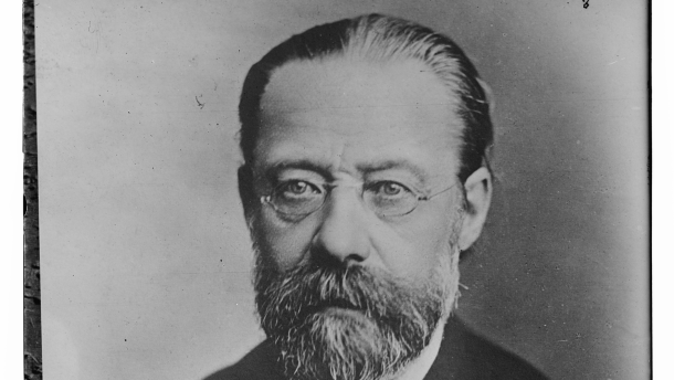 An older man with glasses, a mustache and a beard, dressed in a dark coat with a white shirt, looks toward the camera