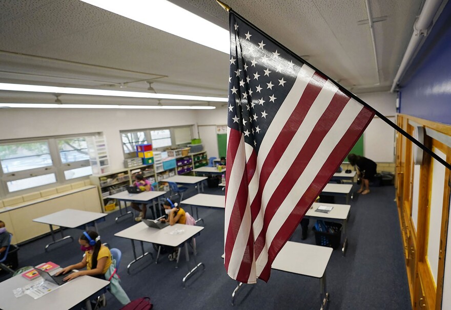 An American flag hangs in a classroom as students work on laptops.