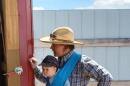 A person in work clothes leans on the door of a barn with a young baby in a sling on their chest.