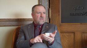 A man wearing a suit sits in an ornately carved wood chair in an office. On the door, written backwards, is, "Chip Neiman."