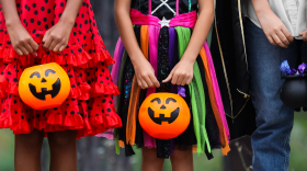 Three children dressed in colorful Halloween costumes hold orange jack-o'-lantern candy buckets, ready for trick-or-treating.