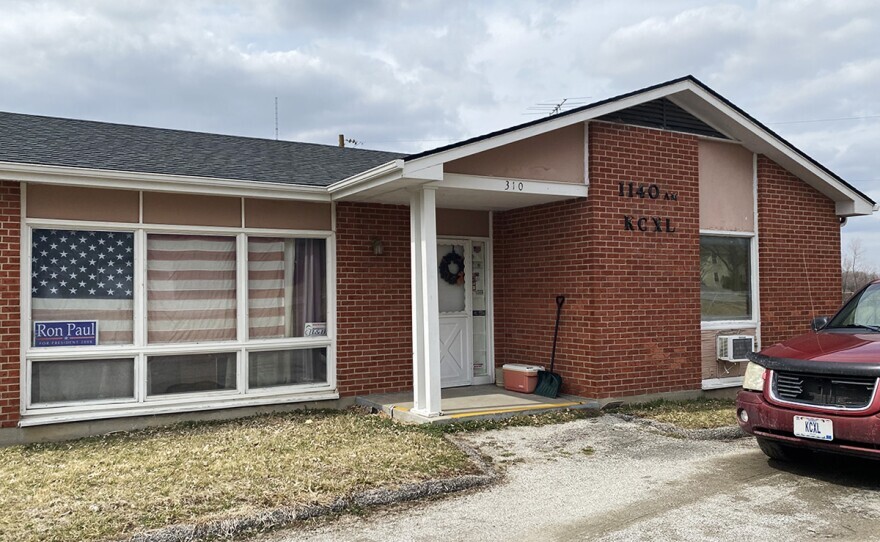 A single-story brick building with the call letters KCXL 1140 am on the side. A faded American flag hangs in the window above a "Ron Paul" campaign sign. A red car with a "KCXL" vanity license plate can be seen in the driveway.