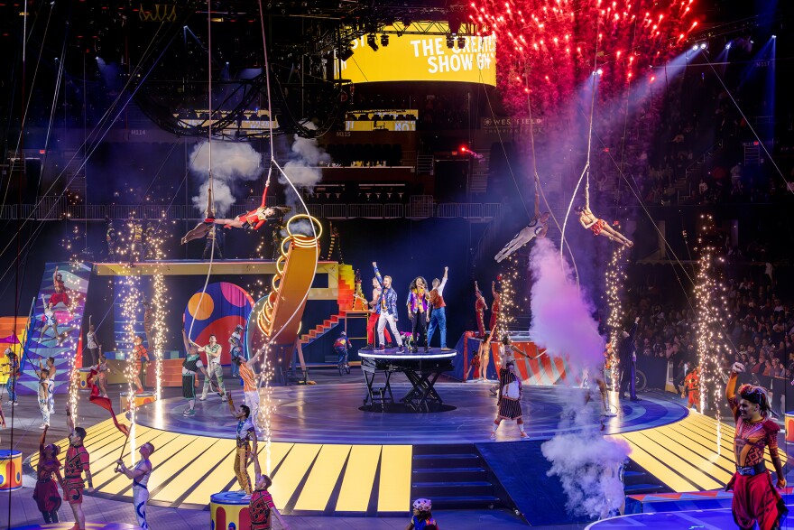 Circus performers on a stage with lights, fireworks, and participants standing and flying in the air. 