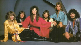 Six band members - two women and four men - sitting on the floor and looking into the camera. All have long hair