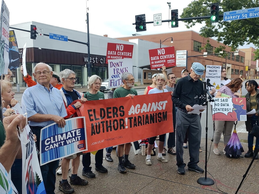 Protesters stand outside behind a man at a microphone. They are holding signs that read, "Elders again authoritarianism" and "Climate can't wait."