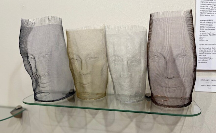 Aluminum wire sculptures of four different heads on a glass shelf and in front of a white wall.