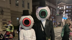 A child and an adult, dressed in tuxedos on top hats an canes, with giant eyeballs as heads, stand in the street at an annual Halloween Parade.