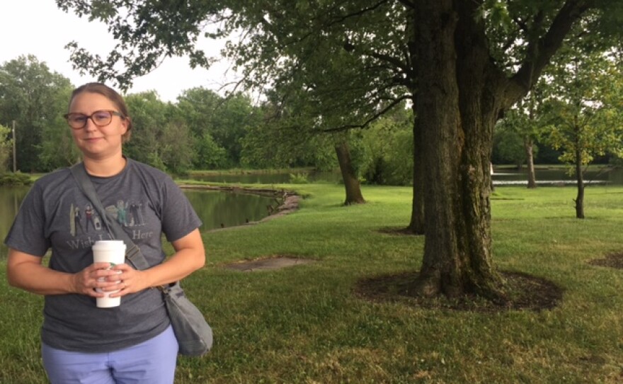 Woman standing with trees, green grass and lake in the background.