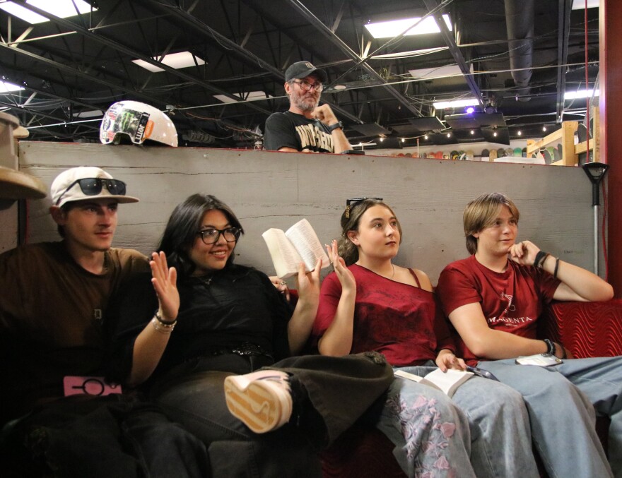 Four youth students sit on a couch. One in black and one in red raise their hands in the air. Behind them is a grey wall and a man leaning on the edge of it. The one student wearing black is holding an open Bible.