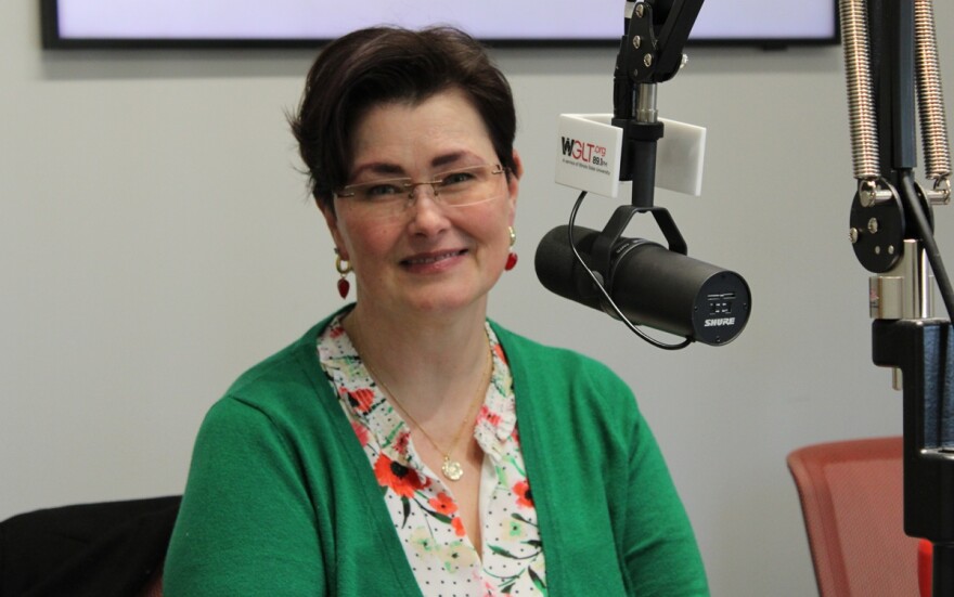 A woman in a green blouse and eyeglasses sits at a desk with a microphone in front of her