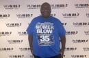 Homer Blow, a long-time radio host in Milwaukee, stands in front of the WUWM 89.7 Milwaukee's NPR photo backdrop wearing a royal blue shirt with the words: "Congratulations Homer Blow Celebrating 35 Years in Radio!"