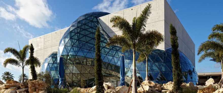 A brown building with a large round glass structure and palm trees in front
